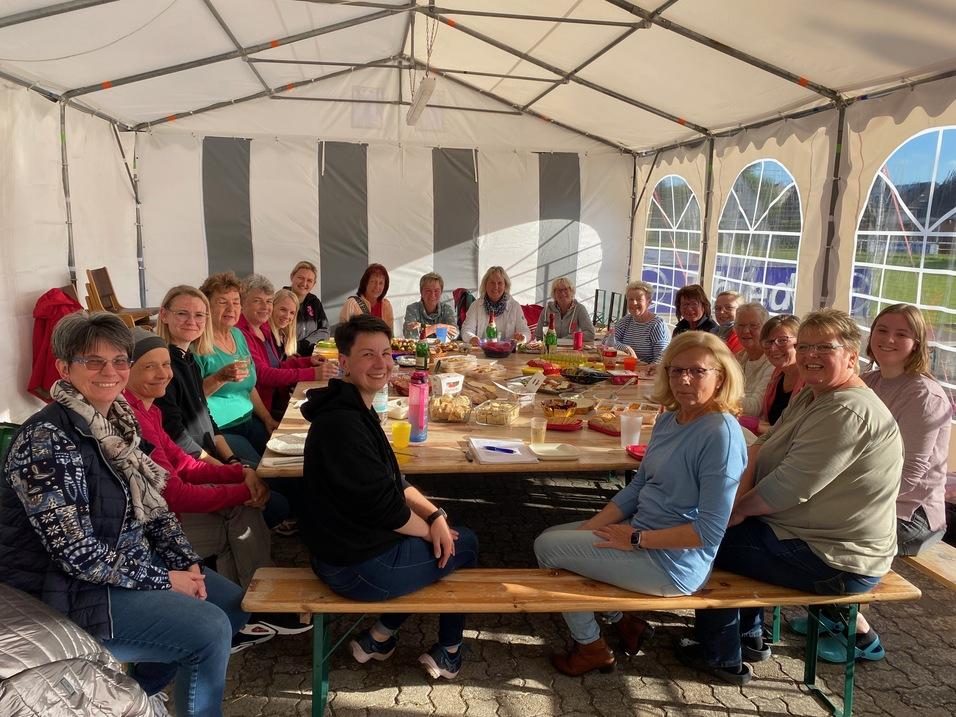 Gruppe von Frauen sitzt um einen Tisch in einem Zelt, umgeben von Speisen und Getr&auml;nken, im Freien.
