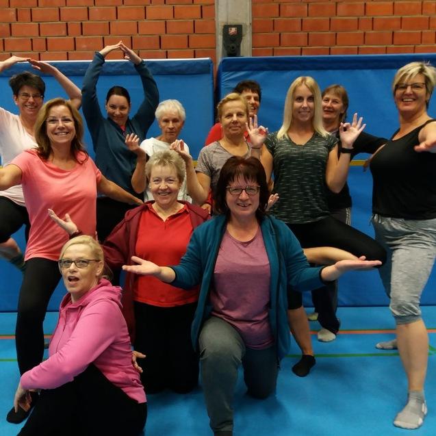 Gruppe von Frauen in Sportkleidung posiert l&auml;chelnd in einem Raum mit blauen Wandmatten und Turnboden.