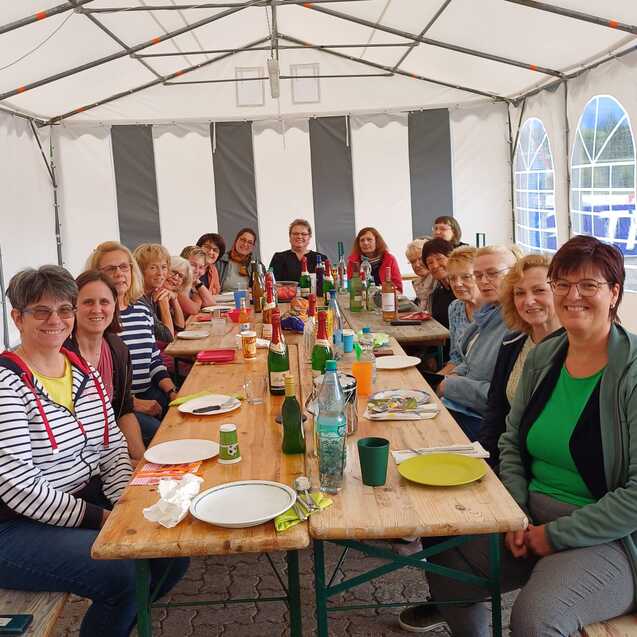 Gruppe von Frauen sitzt an einem langen Tisch unter einem Zelt, umgeben von Getr&auml;nken und Tellern.