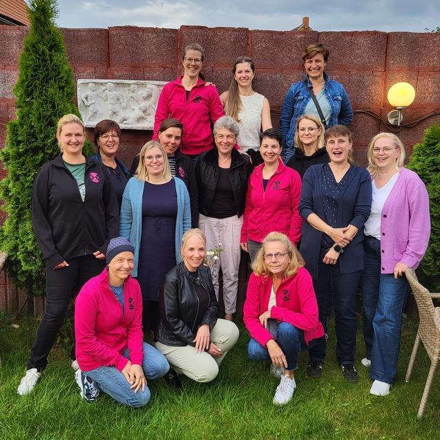 Gruppe von 15 Frauen mit unterschiedlichen Frisuren und Kleidungsstilen, posierend auf einer gr&uuml;nen Wiese vor einer Mauer.