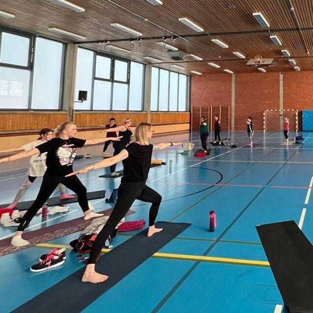 Gruppe von Yogateilnehmern in einem Sportraum, die verschiedene Yoga-Posen auf Matten ausf&uuml;hren.