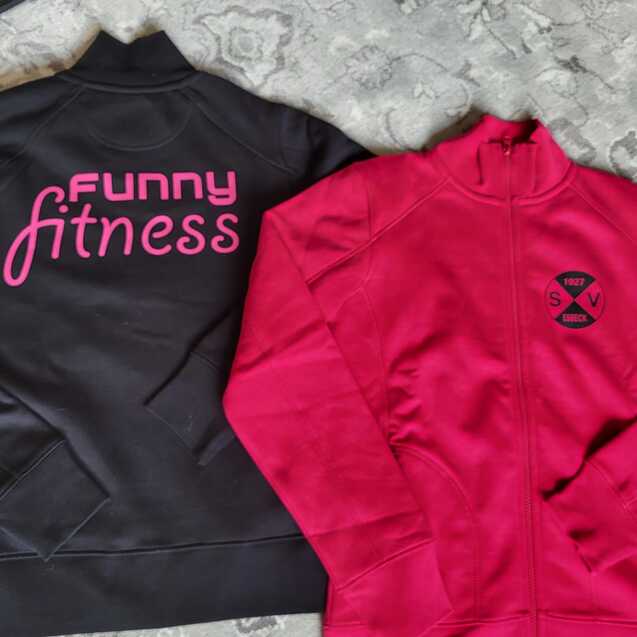 Zwei Sweatjacken liegen auf einem Teppich: links schwarz mit &bdquo;Funny Fitness&ldquo;, rechts pink mit einem Logo.