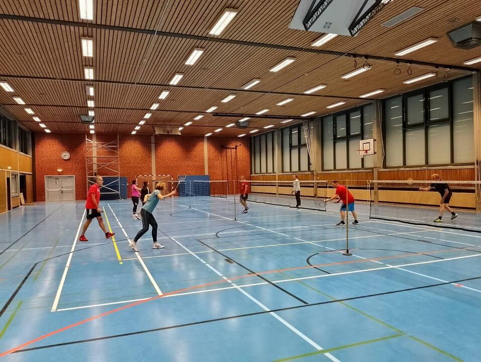 Mehrere Personen spielen Badminton in einer sportlichen Halle mit blauen Bodenmarkierungen und netzen.
