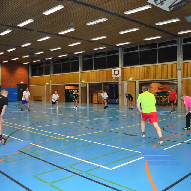 Gruppen von Personen spielen in einer Sporthalle Badminton auf einem blauen Boden mit Linien und Markierungen.
