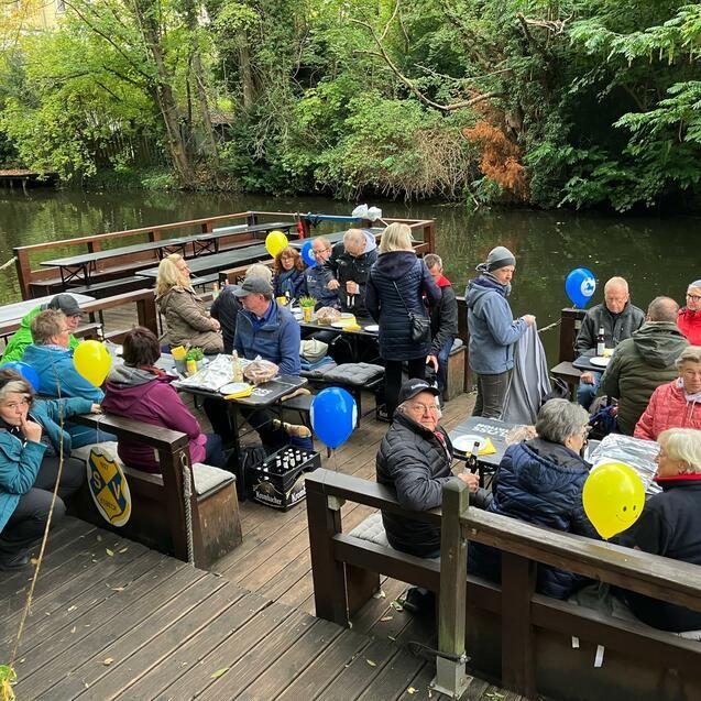 Gruppe von Menschen sitzt an Tischen am Ufer eines Gew&auml;ssers, umgeben von B&auml;umen und bunten Luftballons.