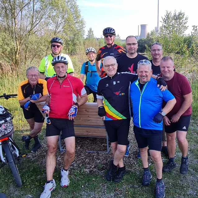 Gruppe von zehn Radfahrern posiert l&auml;chelnd im Freien neben zwei Fahrr&auml;dern vor einer gr&uuml;nen Landschaft.