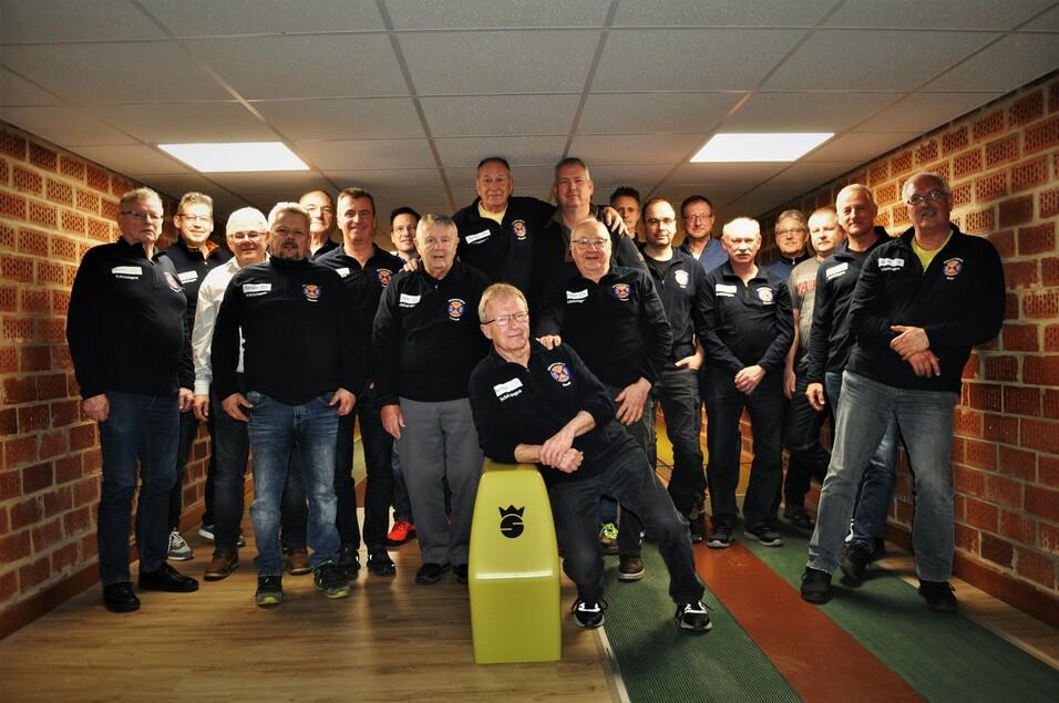 Gruppe von M&auml;nnern in schwarzen Sweatshirts posiert l&auml;chelnd auf einer Bowlingbahn mit einer roten Backsteinwand im Hintergrund.