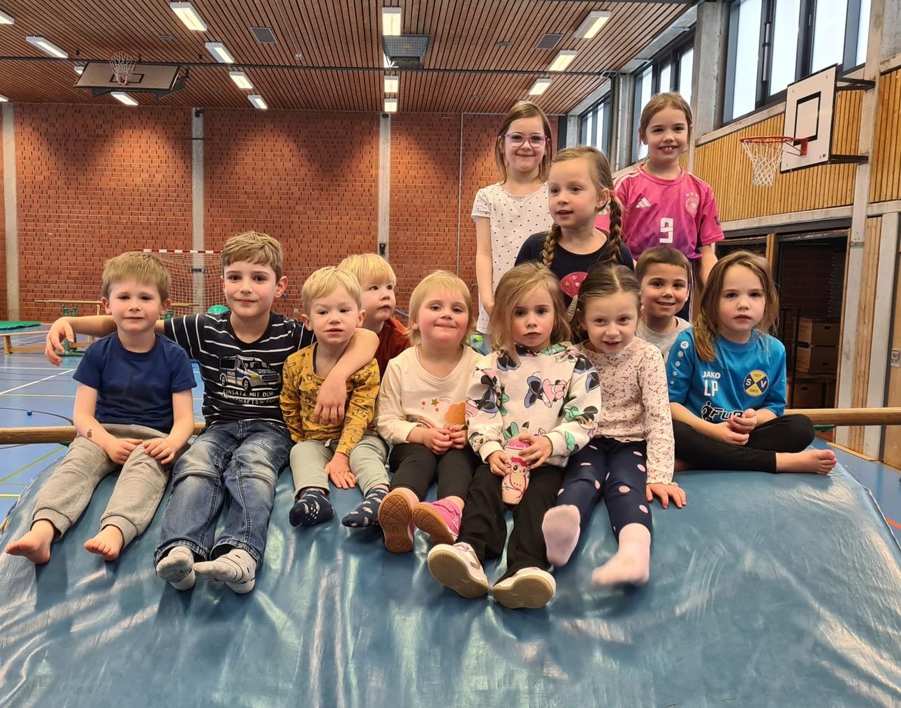 Gruppe von zw&ouml;lf Kindern sitzt auf einem Turnger&auml;t in einer Sporthalle, umgeben von Bewegungsequipment.