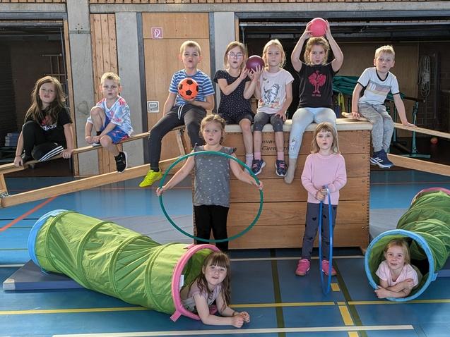 Gruppe von Kindern auf Spielger&auml;ten in einer Turnhalle, einige halten B&auml;lle, andere sitzen in Krabbeltunneln.