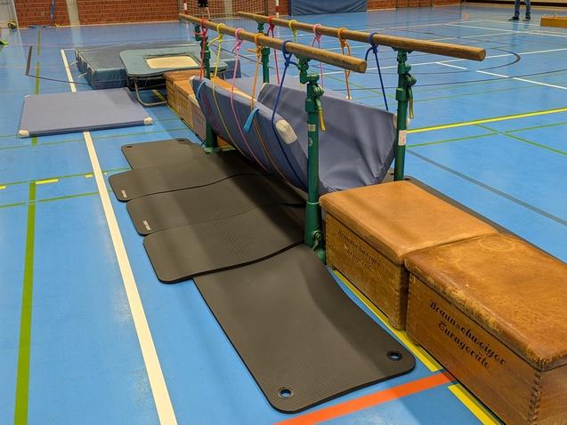 Aufgereihte Turnmatten und B&auml;nke in einer Sporthalle, mit einem Turnger&auml;t aus Holz und Stoffrollen.