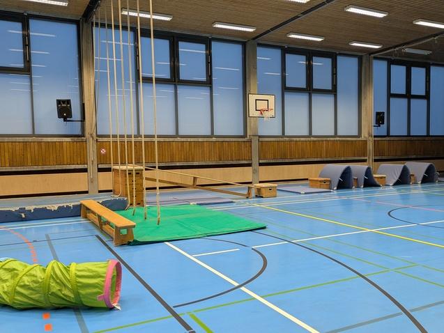 Sporthalle mit blauer Bodenmarkierung, Kletterger&auml;ten, Matten und einem Tunnel. Helle Fenster sorgen f&uuml;r gen&uuml;gend Licht.
