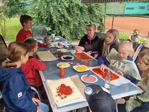 Gruppe von Kindern sitzt an einem Tisch und bereitet Zutaten f&uuml;r Pizzen unter einem Zeltdach vor.