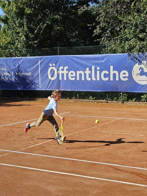 Junge in blauer Shirt und braunen Hosen schl&auml;gt einen Tennisball auf einem Sandplatz w&auml;hrend der Bewegung.