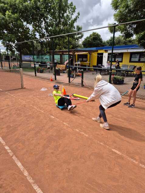 Eine Person sitzt auf dem Tennisplatz und wird von einer anderen mit einem Schl&auml;ger gezogen, w&auml;hrend Kinder im Hintergrund zuschauen.