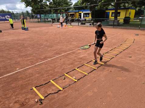 Ein Kind in sportlicher Kleidung &uuml;bt mit einem Tennis-Schl&auml;ger und einer Koordinationsleiter auf einem Sandplatz.