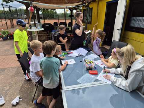 Gruppe von Kindern und einer Betreuerin am Tisch, die kreative Aktivit&auml;ten im Freien durchf&uuml;hren. Tennisplatz im Hintergrund.