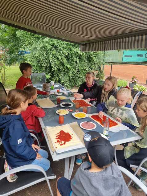 Gruppe von Kindern sitzt an einem Tisch und bereitet Zutaten f&uuml;r Pizzen unter einem Zeltdach vor.