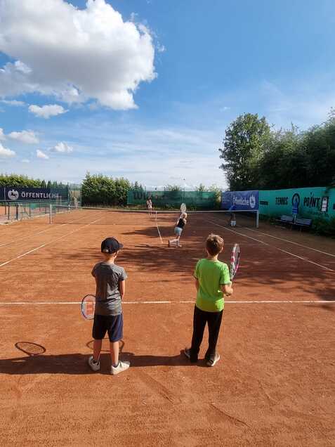 Zwei Kinder stehen mit Tennisschl&auml;gern auf einem Tennisplatz, w&auml;hrend ein weiteres Kind im Hintergrund spielt.
