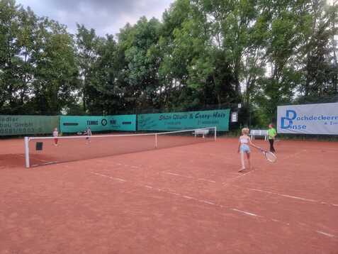Zwei Kinder spielen Tennis auf einem roten Sandplatz, umgeben von gr&uuml;nen B&auml;umen und Werbebannern.