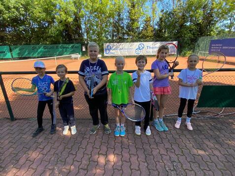 Gruppe von acht Kindern mit Tennisschl&auml;gern auf einem Tennisplatz, umgeben von gr&uuml;nen B&auml;umen und einem Netz.