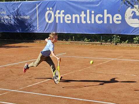 Junge in blauer Shirt und braunen Hosen schl&auml;gt einen Tennisball auf einem Sandplatz w&auml;hrend der Bewegung.