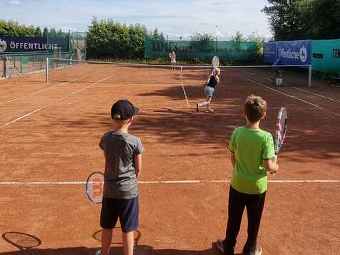 Zwei Kinder stehen mit Tennisschl&auml;gern auf einem Tennisplatz, w&auml;hrend ein weiteres Kind im Hintergrund spielt.