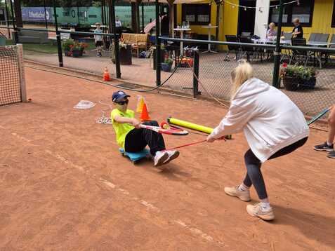 Eine Person sitzt auf dem Tennisplatz und wird von einer anderen mit einem Schl&auml;ger gezogen, w&auml;hrend Kinder im Hintergrund zuschauen.