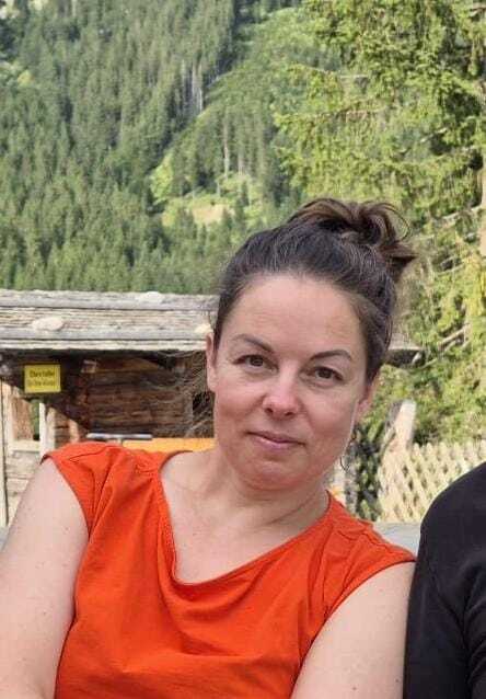 Frau mit Dutt tr&auml;gt ein oranges Oberteil, sitzt vor alpenl&auml;ndischer Landschaft mit B&auml;umen und H&uuml;tten im Hintergrund.