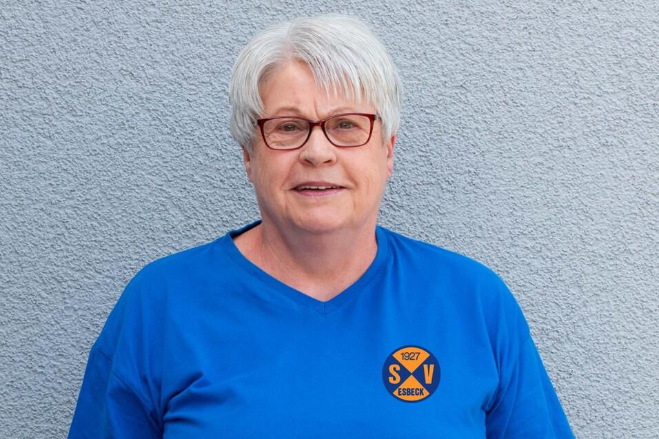 Portr&auml;t einer l&auml;chelnden Frau mit grau-wei&szlig;en Haaren in einem blauen T-Shirt vor einer grauen Wand.