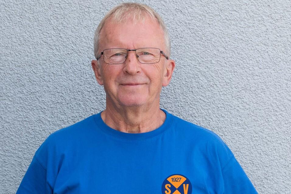 &Auml;lterer Mann mit Brille tr&auml;gt ein blaues T-Shirt mit dem Logo eines Sportvereins vor einer grauen Wand.