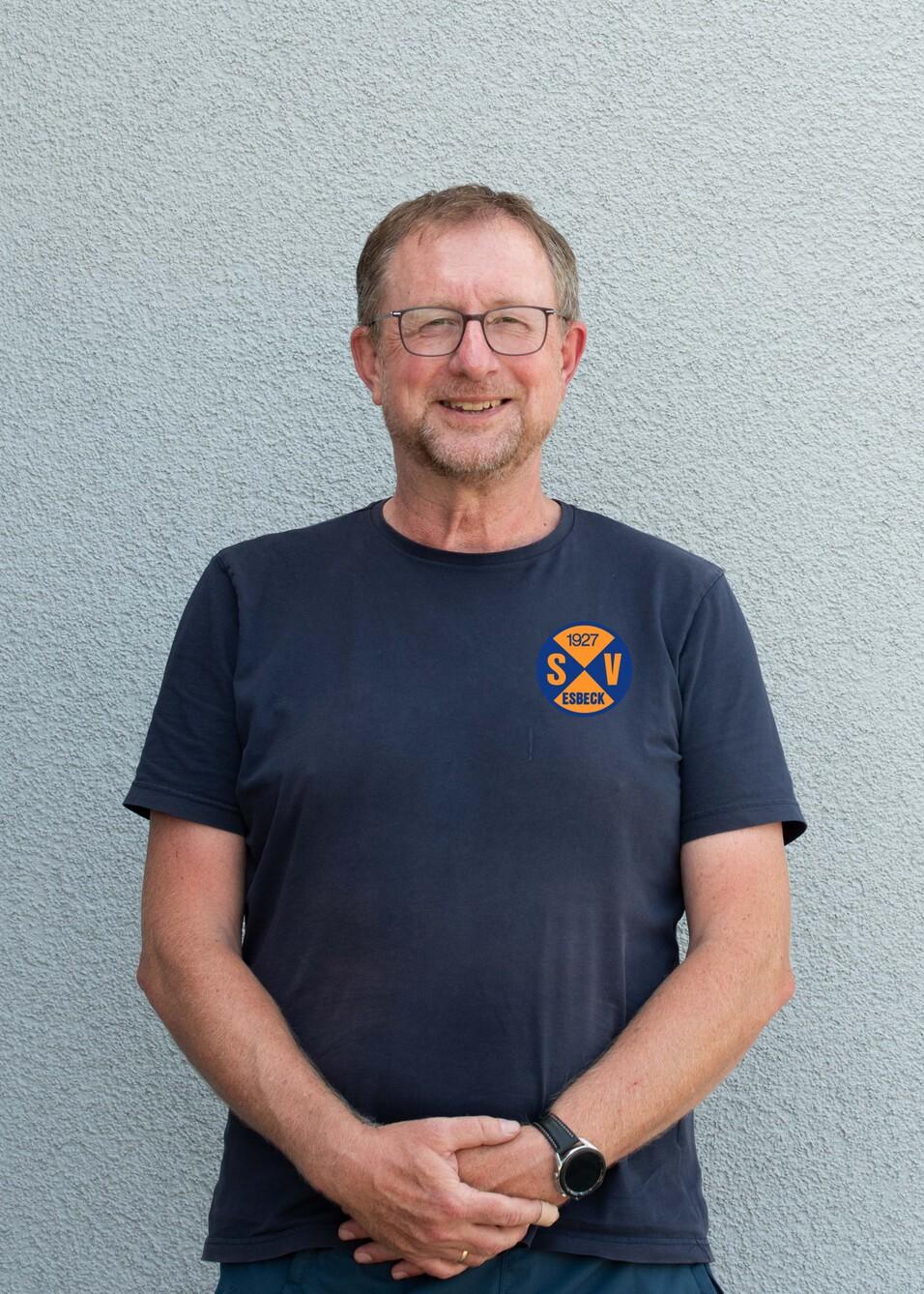 Portr&auml;t eines l&auml;chelnden Mannes mit Brille, der in einem T-Shirt mit Vereinslogo vor einer grauen Wand steht.