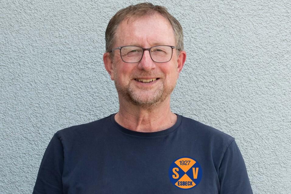 Portr&auml;t eines l&auml;chelnden Mannes mit Brille, der in einem T-Shirt mit Vereinslogo vor einer grauen Wand steht.