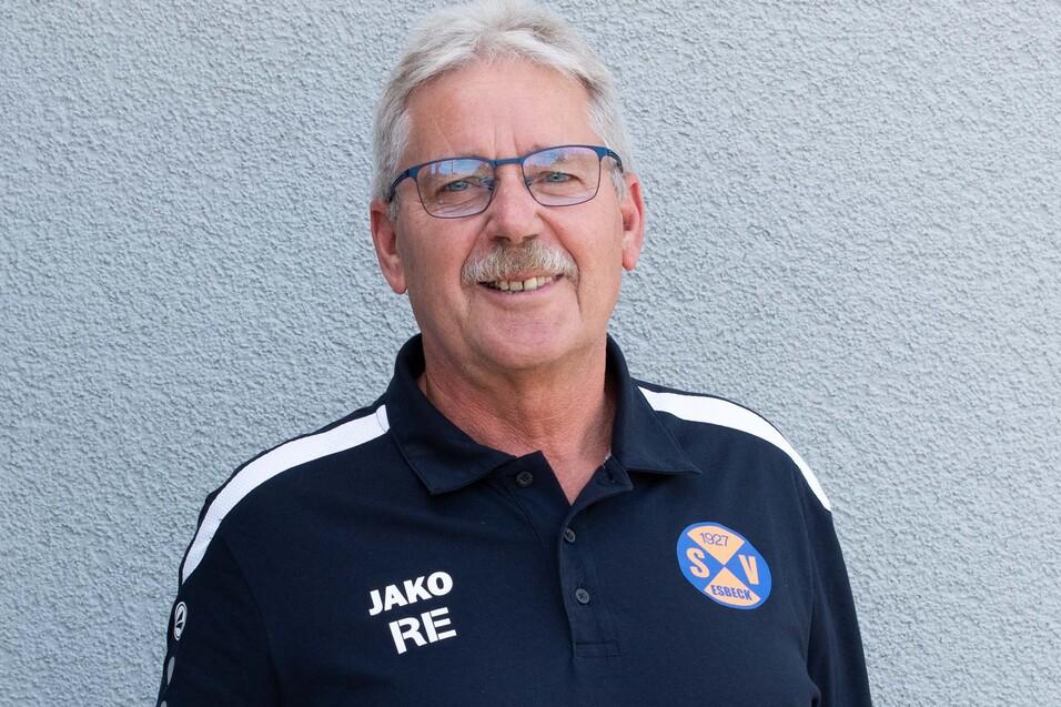 M&auml;nnlicher Trainer in einem schwarzen Poloshirt mit Vereinslogo steht vor einer grauen Wand.