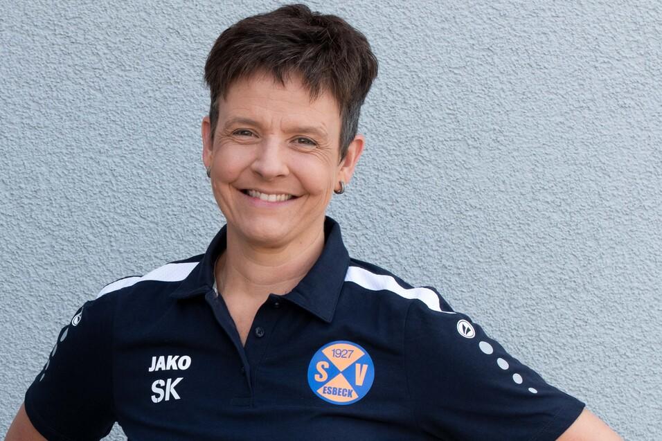 Frau mit kurzem Haar tr&auml;gt ein dunkles Sportshirt mit Vereinslogo und steht l&auml;chelnd vor einer grauen Wand.