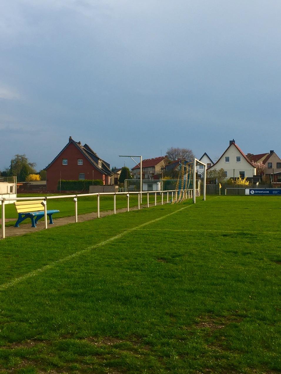 Gr&uuml;ner Fu&szlig;ballplatz mit Tor und Bank, umgeben von Wohnh&auml;usern im Hintergrund unter bew&ouml;lktem Himmel.