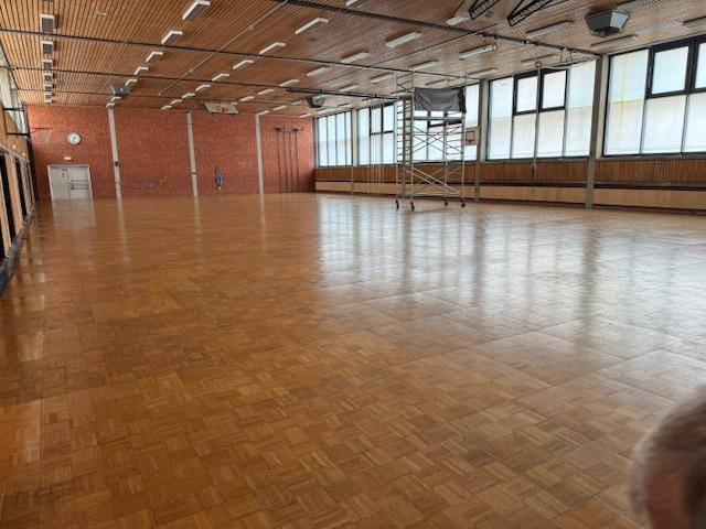 Leere Turnhalle mit Holzfu&szlig;boden, gro&szlig;en Fenstern und einem Bauger&uuml;st in der Mitte.