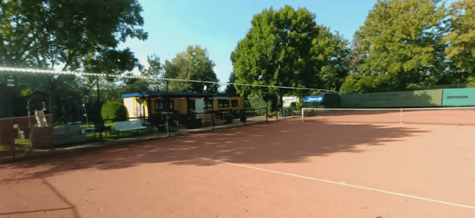 Blick auf einen Sandtennisplatz mit einer gelben Umkleidekabine und gr&uuml;nen B&auml;umen im Hintergrund.