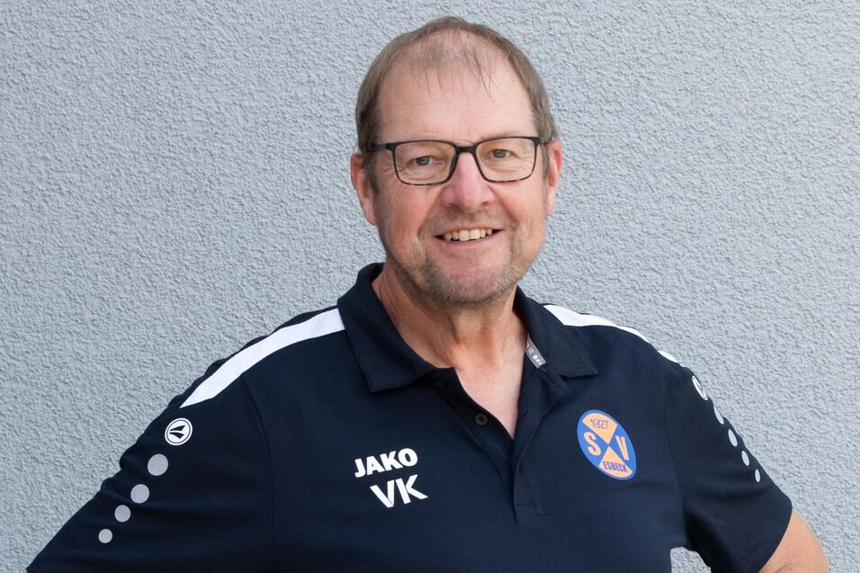 Portrait eines l&auml;chelnden Mannes in einem schwarzen Sporttrikot mit Brille vor grauem Hintergrund.