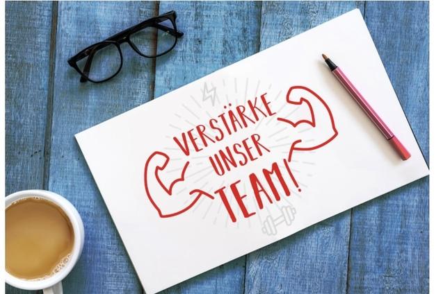 Wei&szlig;es Blatt mit der Aufschrift &bdquo;Verst&auml;rke unser Team!&ldquo; und Grafik von muskul&ouml;sen Armen auf einem blauen Tisch. Gl&auml;ser und Tasse Kaffee.
