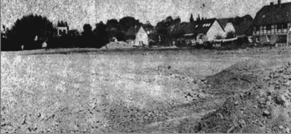 Schwarz-Wei&szlig;-Foto einer leeren, unebenen Fl&auml;che mit Geb&auml;uden im Hintergrund und B&auml;umen am Rand.