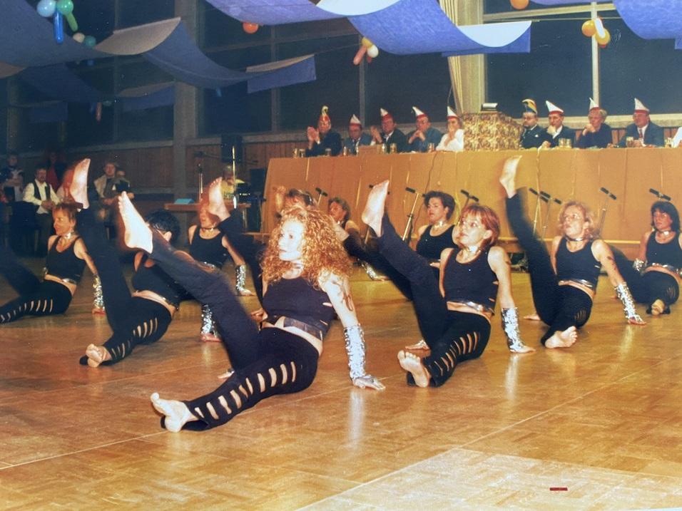 Tanzgruppe in Schwarz f&uuml;hrt synchronisierte Bewegungen auf einer B&uuml;hne mit Zuschauern im Hintergrund aus.