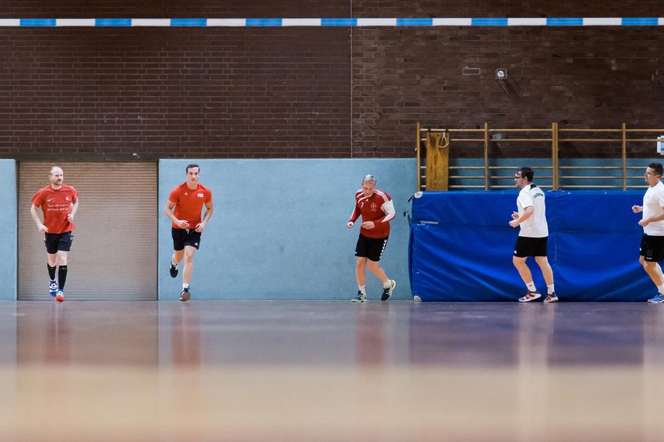 Fünf Spieler in Sportkleidung trainieren in einer Sporthalle, mit einem Tor und blauen Wänden im Hintergrund.