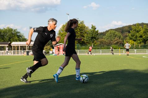 Mann und Mädchen laufen auf einem Fußballfeld, während der Ball auf dem Rasen liegt. Im Hintergrund sind weitere Spieler.