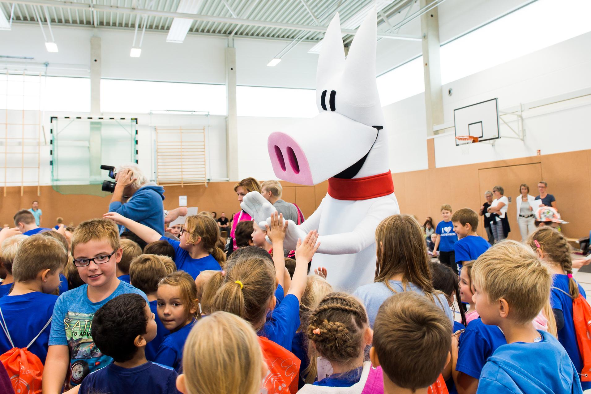 Ein großer, weißer Stofftier-Charakter interagiert mit aufgeregten Kindern in blauen T-Shirts in einer Turnhalle.