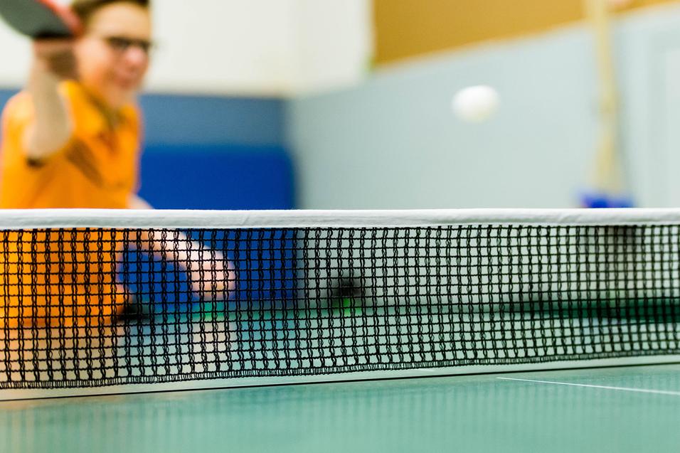 Tischtennisschläger in der Hand eines Spielers, im Hintergrund ein Ball über das Netz fliegend auf einer grünen Tischtennisplatte.